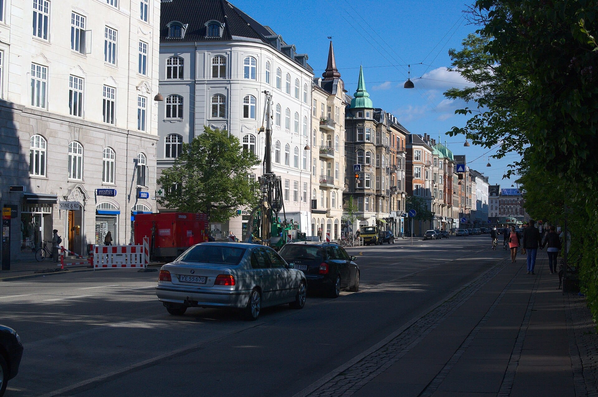 ถนนบูเลอวาร์ดที่มีต้นไม้เรียงรายใน Østerbro เมือง Copenhagen พร้อมอาคารประวัติศาสตร์หลายชั้น รถยนต์จอดอยู่ริมถนน และท้องฟ้าสีฟ้าแจ่มใส