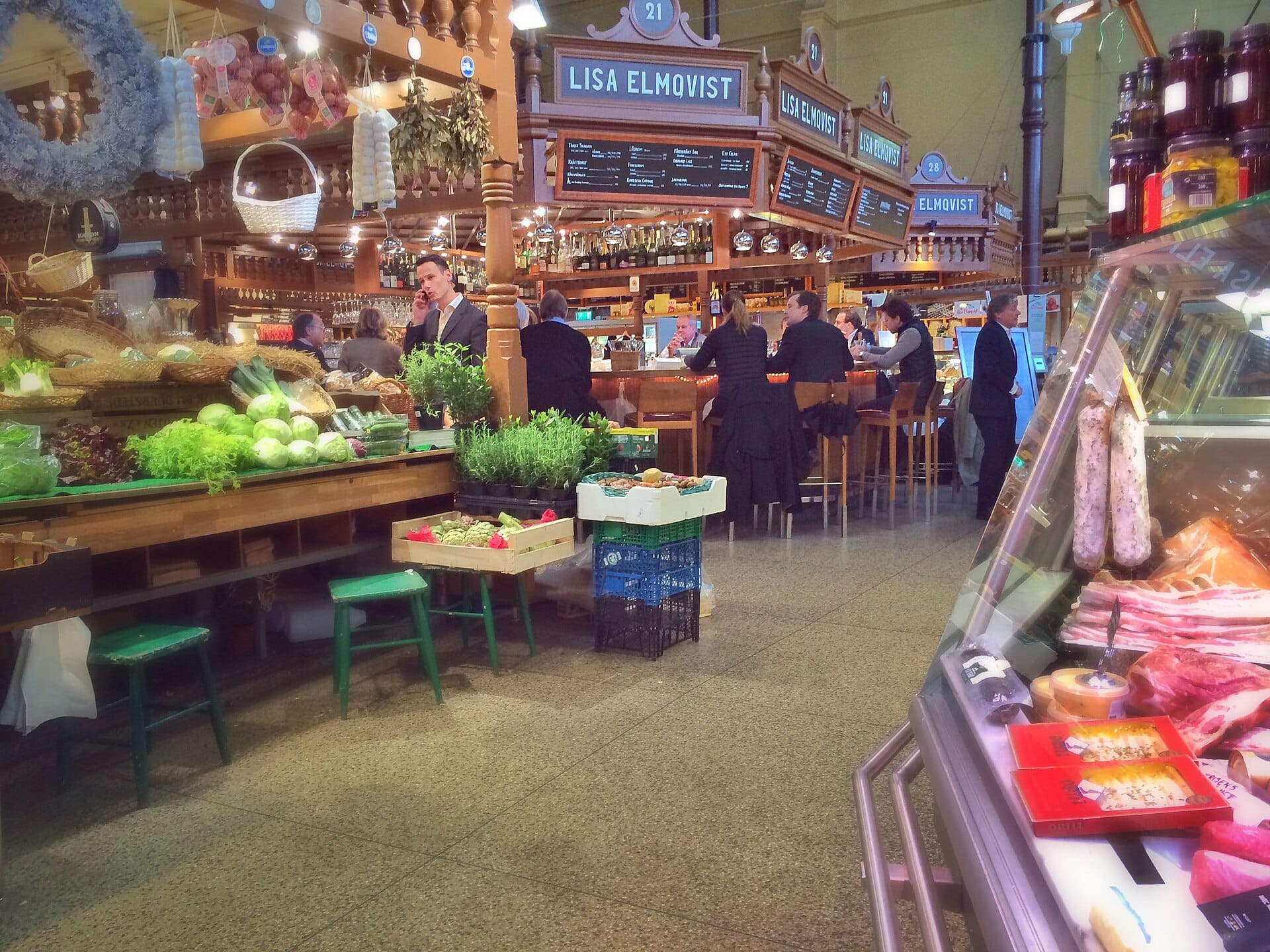 บรรยากาศภายใน Östermalm Food Hall มีผักสด เคาน์เตอร์เดลิ และคนกำลังกินอาหารที่บาร์ร้าน Lisa Elmqvist ใต้ป้ายไม้คลาสสิก