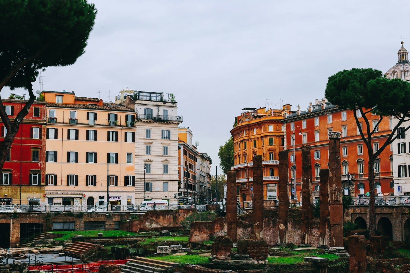 View of Ostia Antica ruins with ancient stone columns in the foreground, surrounded by colorful old buildings and pine trees under a cloudy sky.