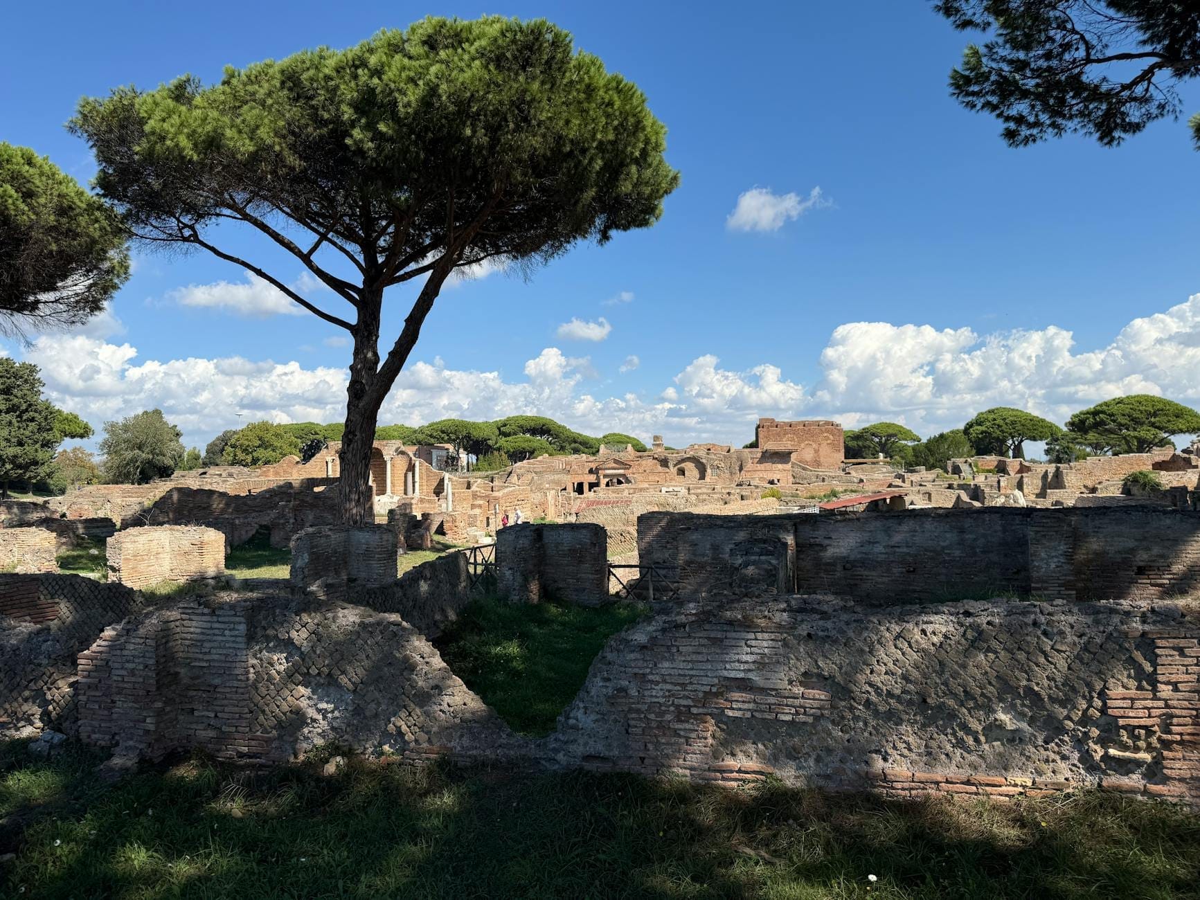 Ruins and ancient brick walls of Ostia Antica under blue sky with pine trees, showing the archaeological site’s open and expansive layout.