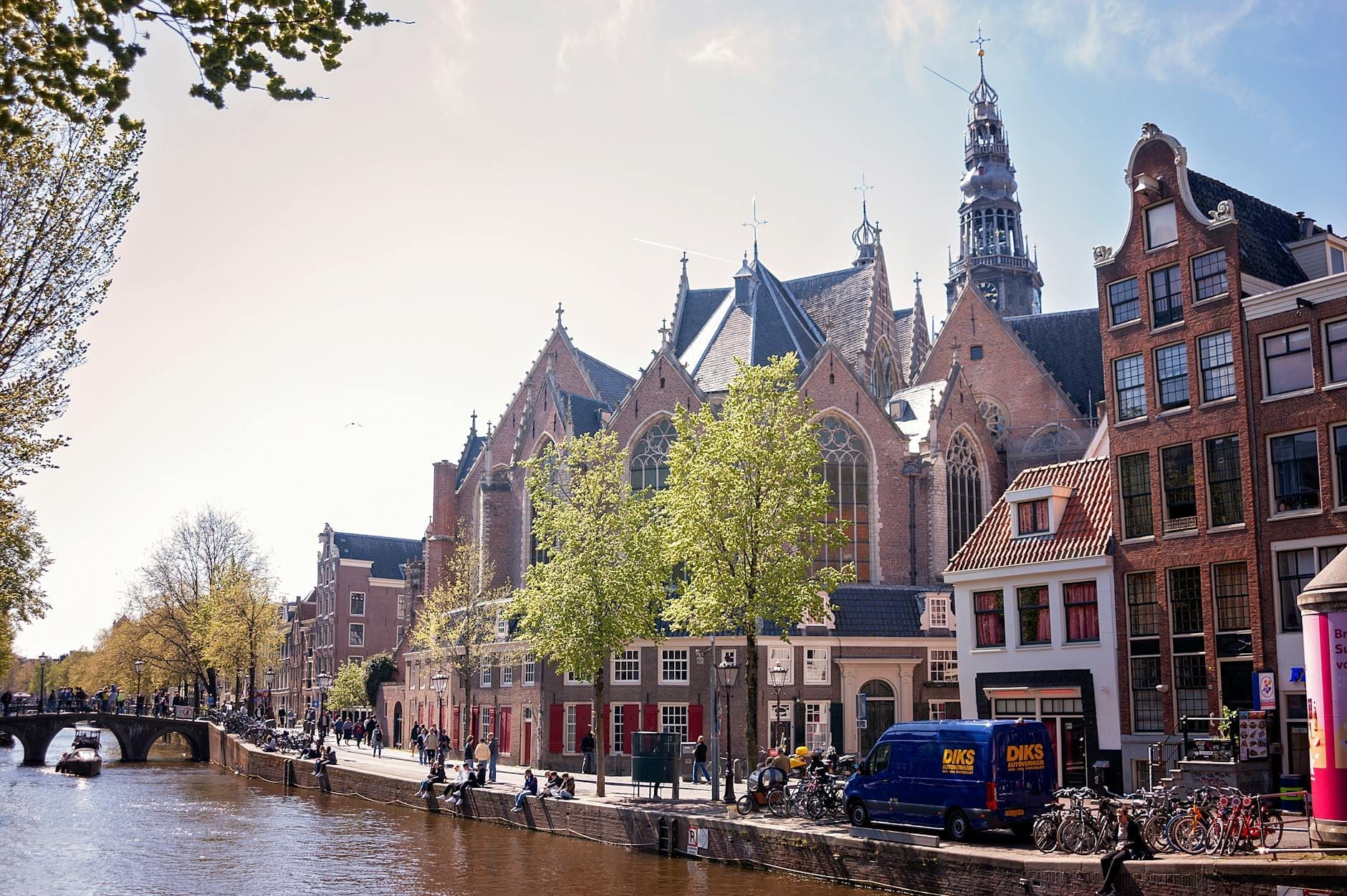 Vista de la arquitectura gótica de la Oude Kerk junto a un canal de Ámsterdam, rodeada de edificios históricos y calles arboladas bajo un cielo despejado.