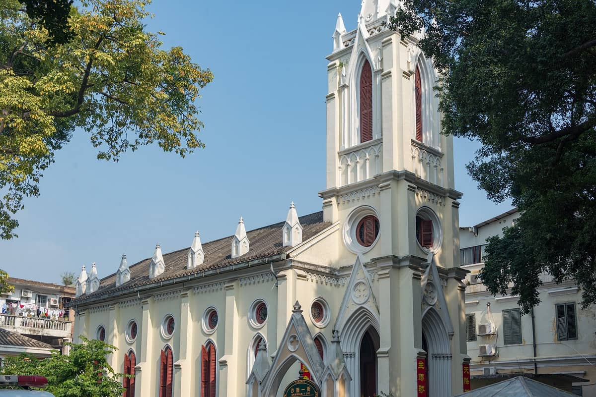 Landscape view of the yellow Gothic-style Our Lady of Lourdes Chapel on Shamian Island, Guangzhou, with pointed arches, red window shutters, and trees framing the chapel against a blue sky.