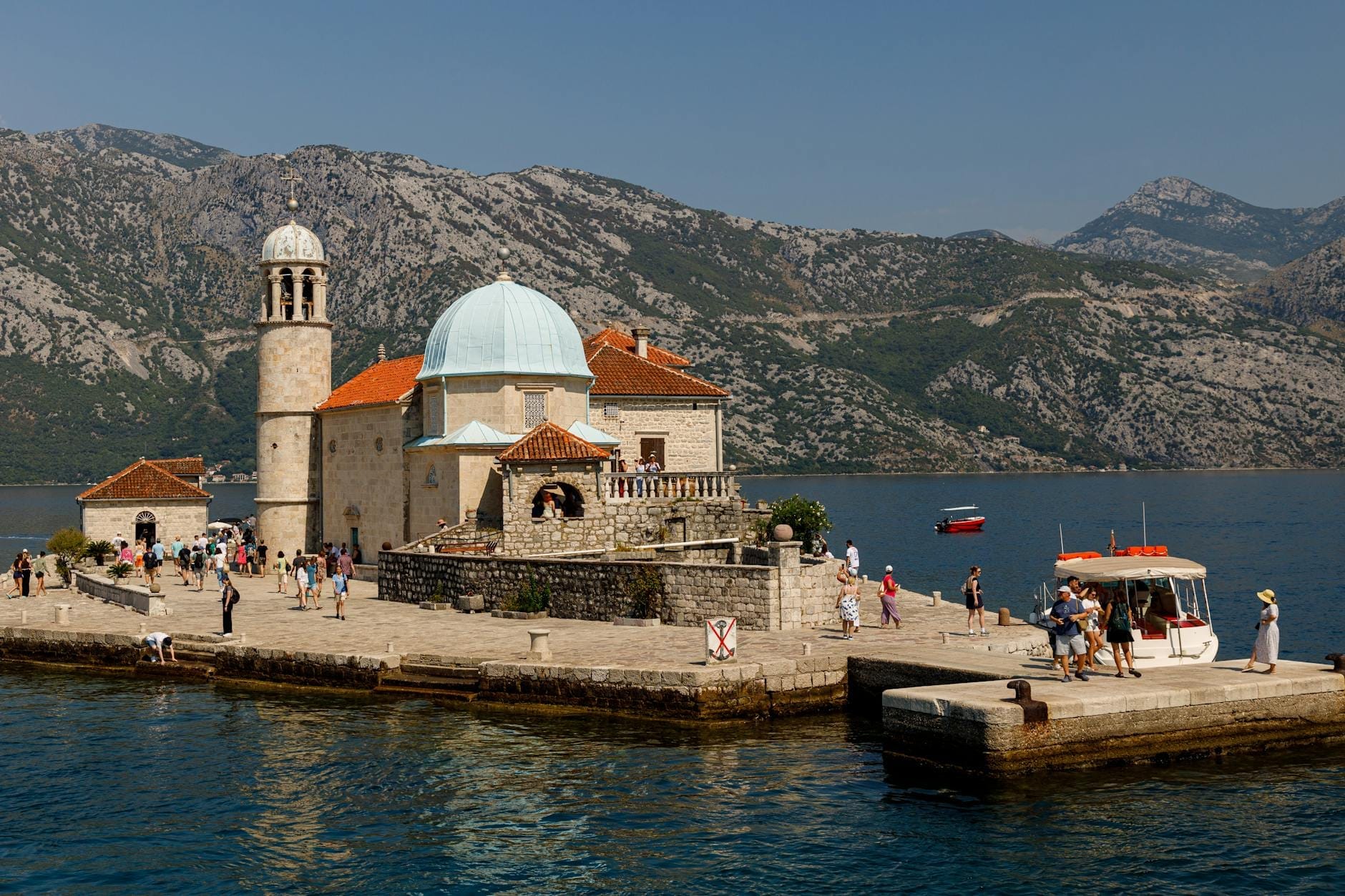 La chiesa della Madonna dello Scoglio con la cupola azzurra e il campanile in pietra su un'isola artificiale, circondata da turisti, barche e montagne imponenti nella Baia di Kotor.