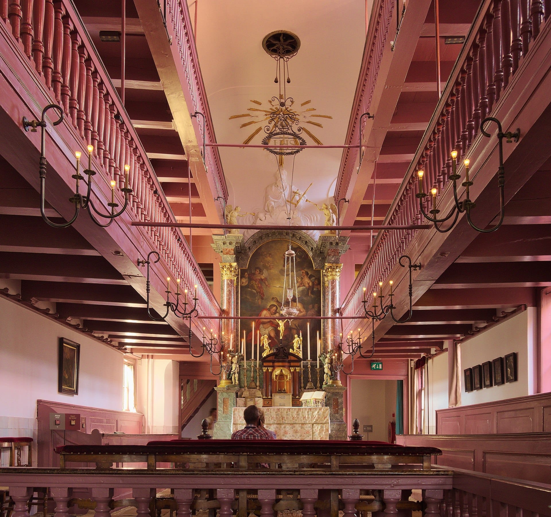 Vista interior de la iglesia del Museo Ons' Lieve Heer op Solder, con un altar ornamentado, balcones de madera rosa, lámparas de araña y un visitante sentado.