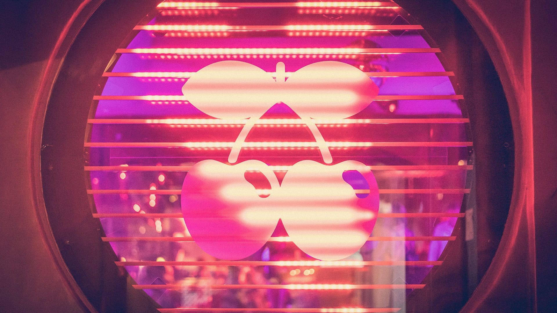 L'iconico logo della ciliegia di Pacha Ibiza illuminato dalle luci rosa e viola del club, visto attraverso le veneziane orizzontali all'ingresso.