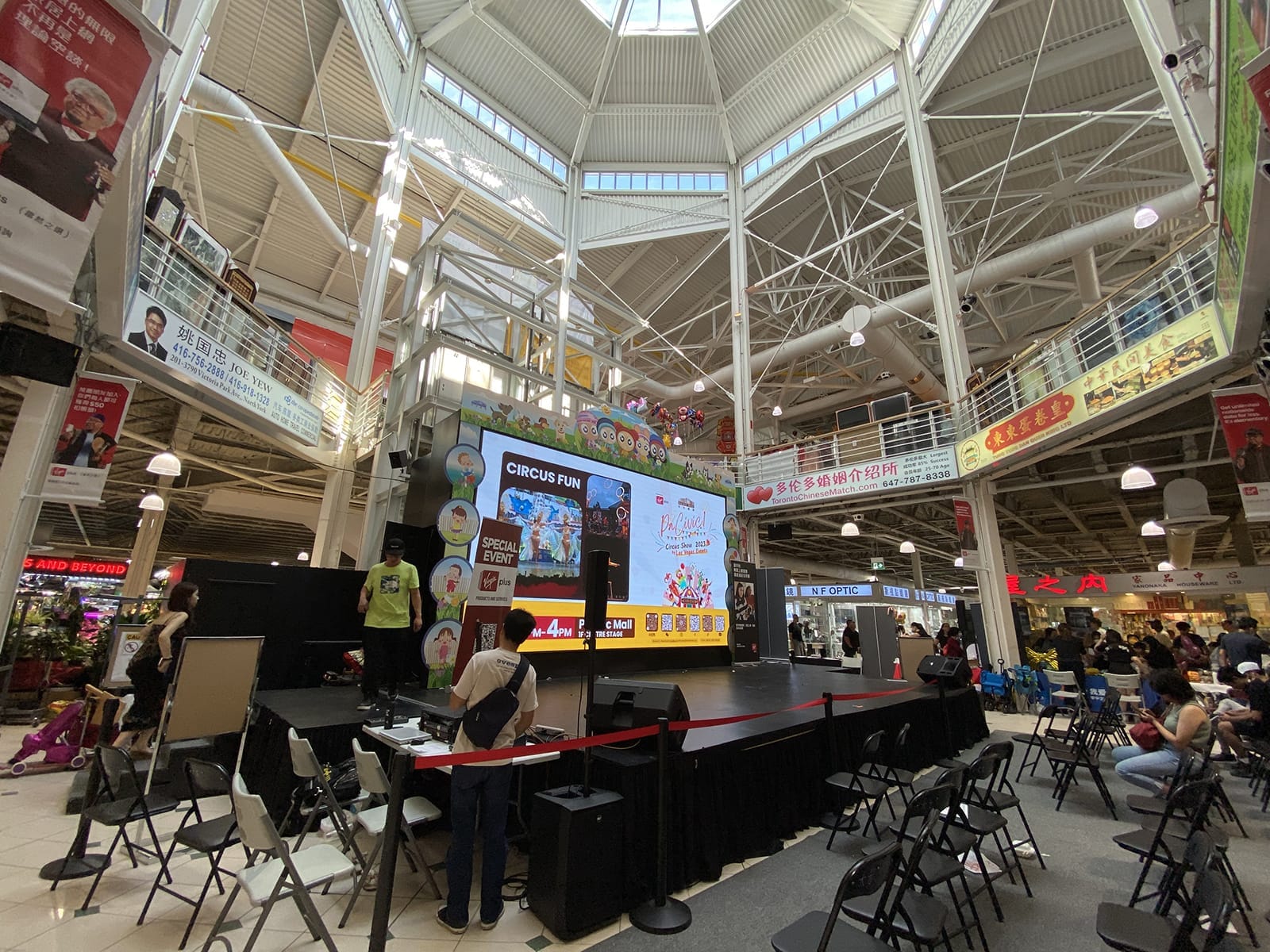 มุมมองกว้างของโถงกลาง Pacific Mall ที่สว่างไสว พร้อมเวที จอดิจิทัลแสดงกิจกรรม เก้าอี้ และผู้คนใต้หลังคากระจก