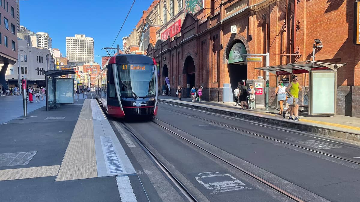 Il tram leggero di Sydney davanti all'ingresso del Market City, dove ha sede il Paddy's Markets di Haymarket, con persone che camminano e il cartello del mercato ben visibile.