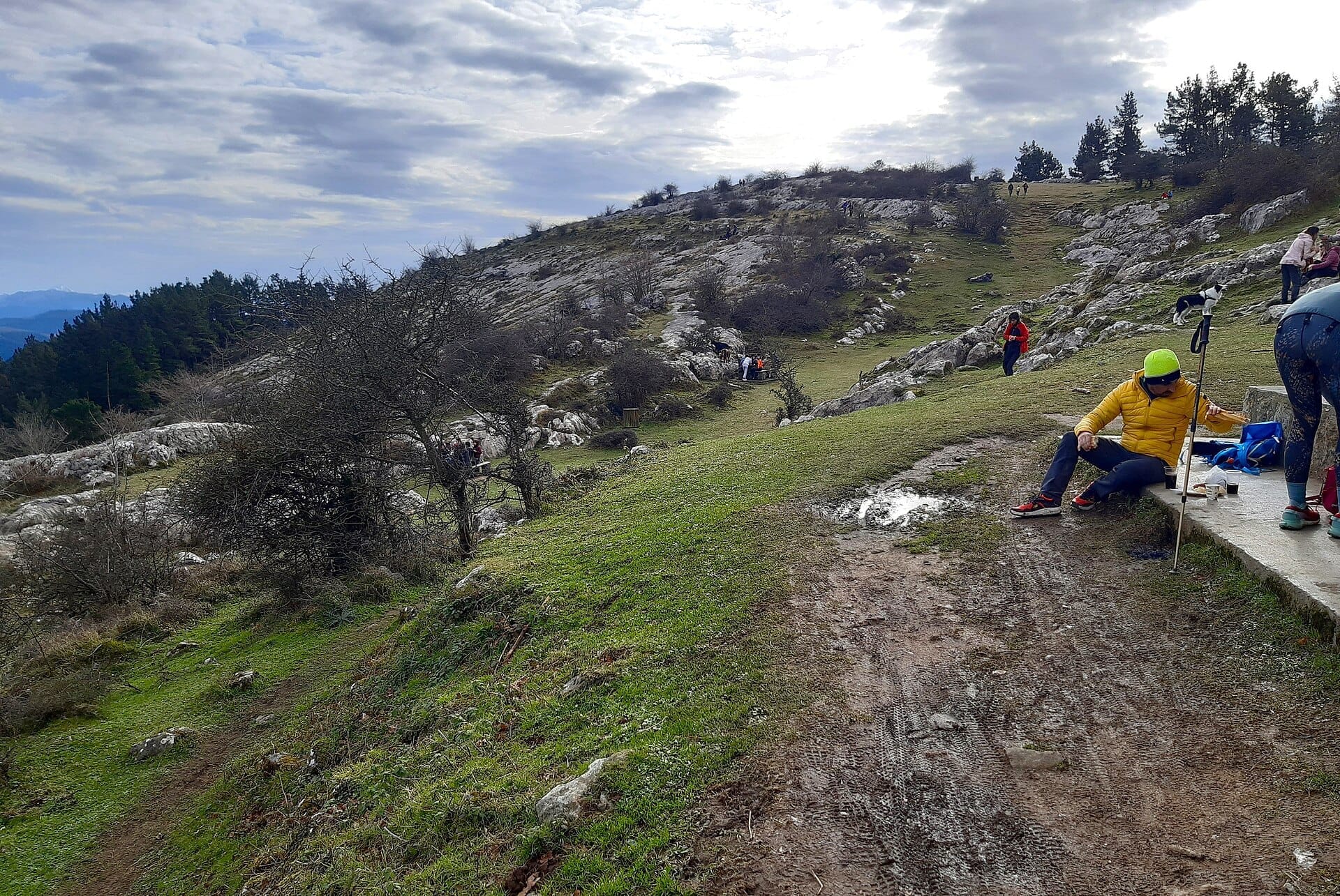 Pagasarri Mountain hike trail with rocky slopes, scattered trees, and several people enjoying the outdoors under a cloudy sky near Bilbao.