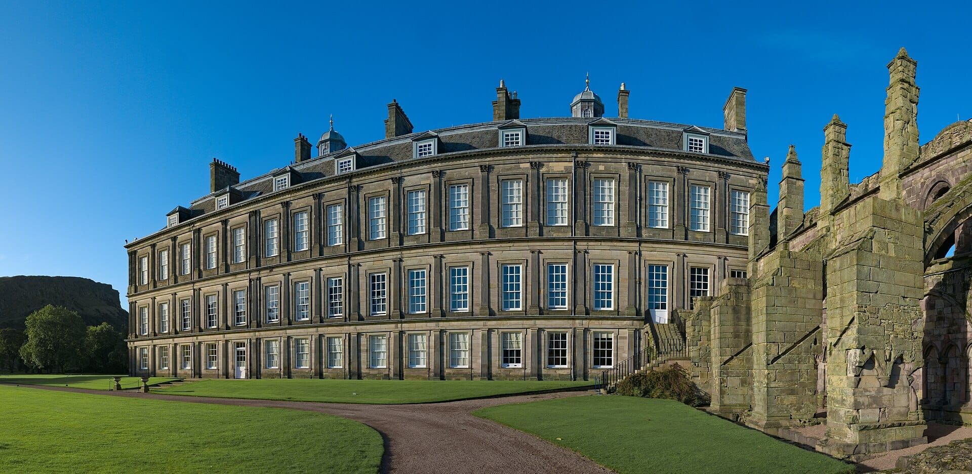 Weitwinkelansicht des Palace of Holyroodhouse mit seiner geschwungenen Fassade und den angrenzenden Ruinen der Holyrood Abbey im hellen Sonnenlicht vor strahlend blauem Himmel.