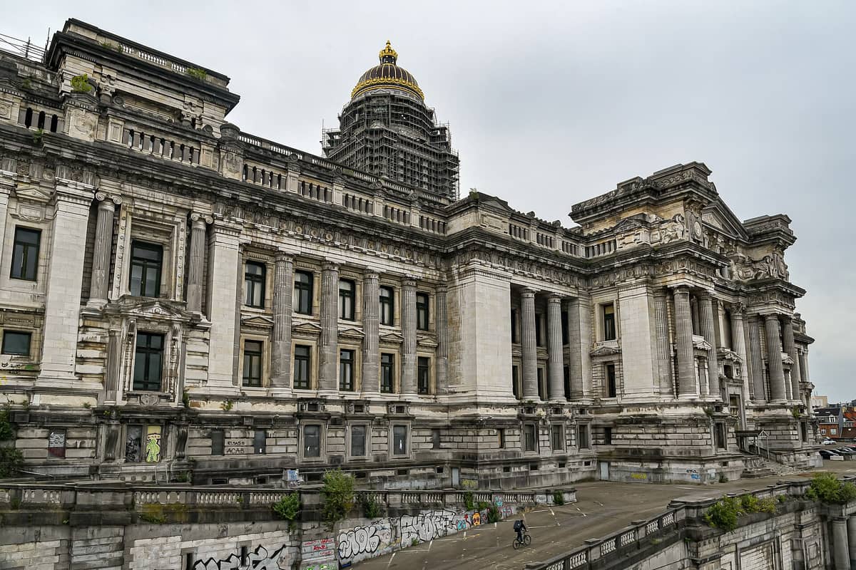 Vista externa ampla do neoclássico Palácio de Justiça de Bruxelas, mostrando sua fachada imponente, colunas e cúpula coberta por andaimes.