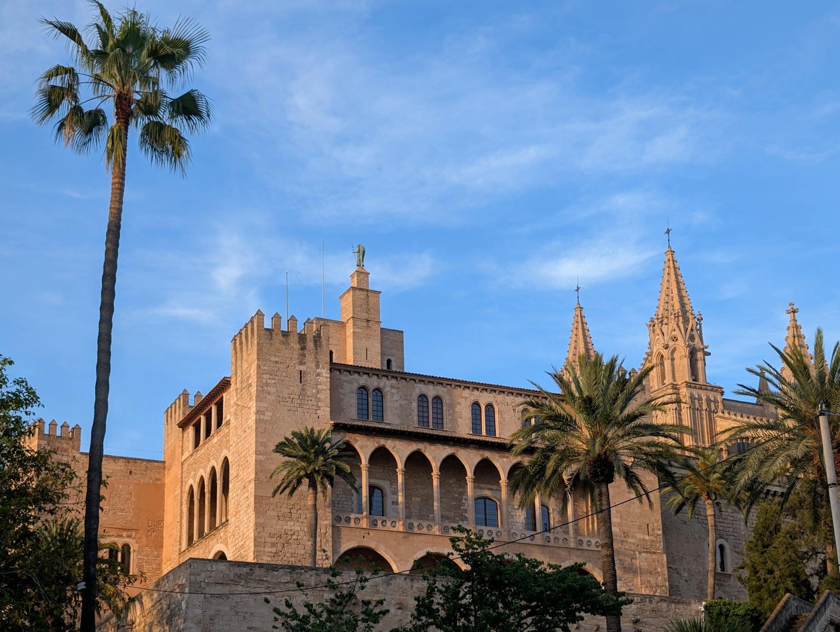 Il Palazzo de La Almudaina a Palma, Mallorca, è raffigurato con le sue mura in pietra, i merli e le palme sotto un cielo azzurro limpido.