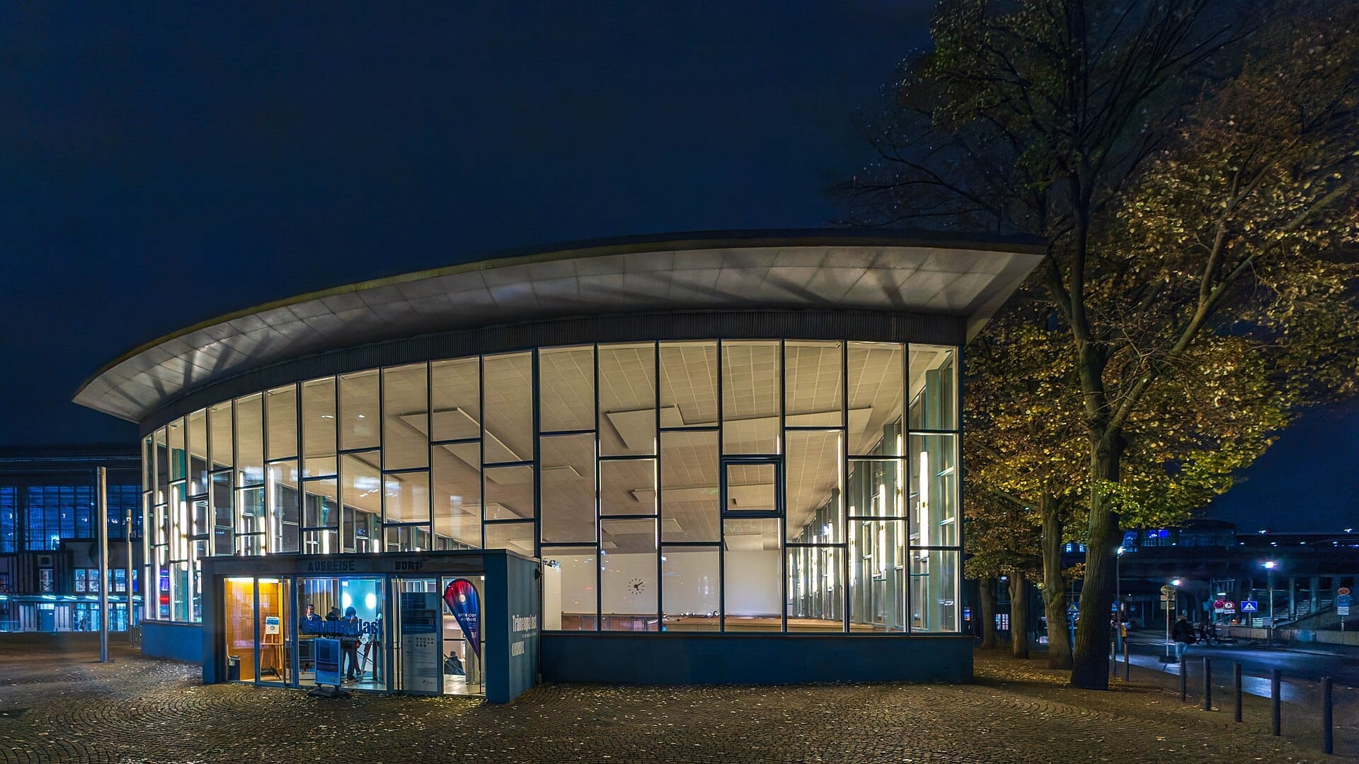 Il Palazzo delle Lacrime a Berlino illuminato di notte, con la sua facciata in vetro, l'architettura moderna e gli alberi autunnali circostanti.