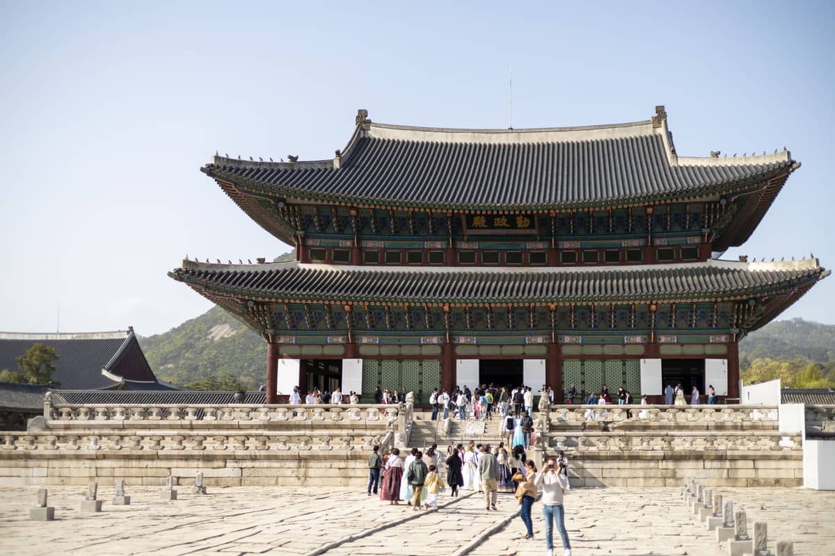 Grande grupo de visitantes no Salão Geunjeongjeon no Palácio Gyeongbokgung, com o majestoso salão tradicional de madeira de dois andares e as montanhas ao fundo.