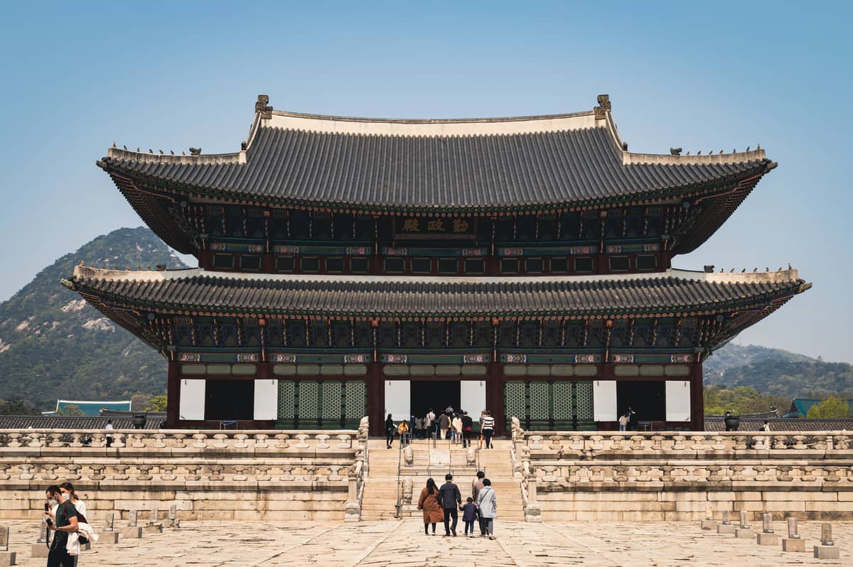 Vista ampla do salão do trono principal do Palácio Gyeongbokgung em Seul, com pessoas caminhando e um céu azul claro sobre o marco arquitetônico da era Joseon.