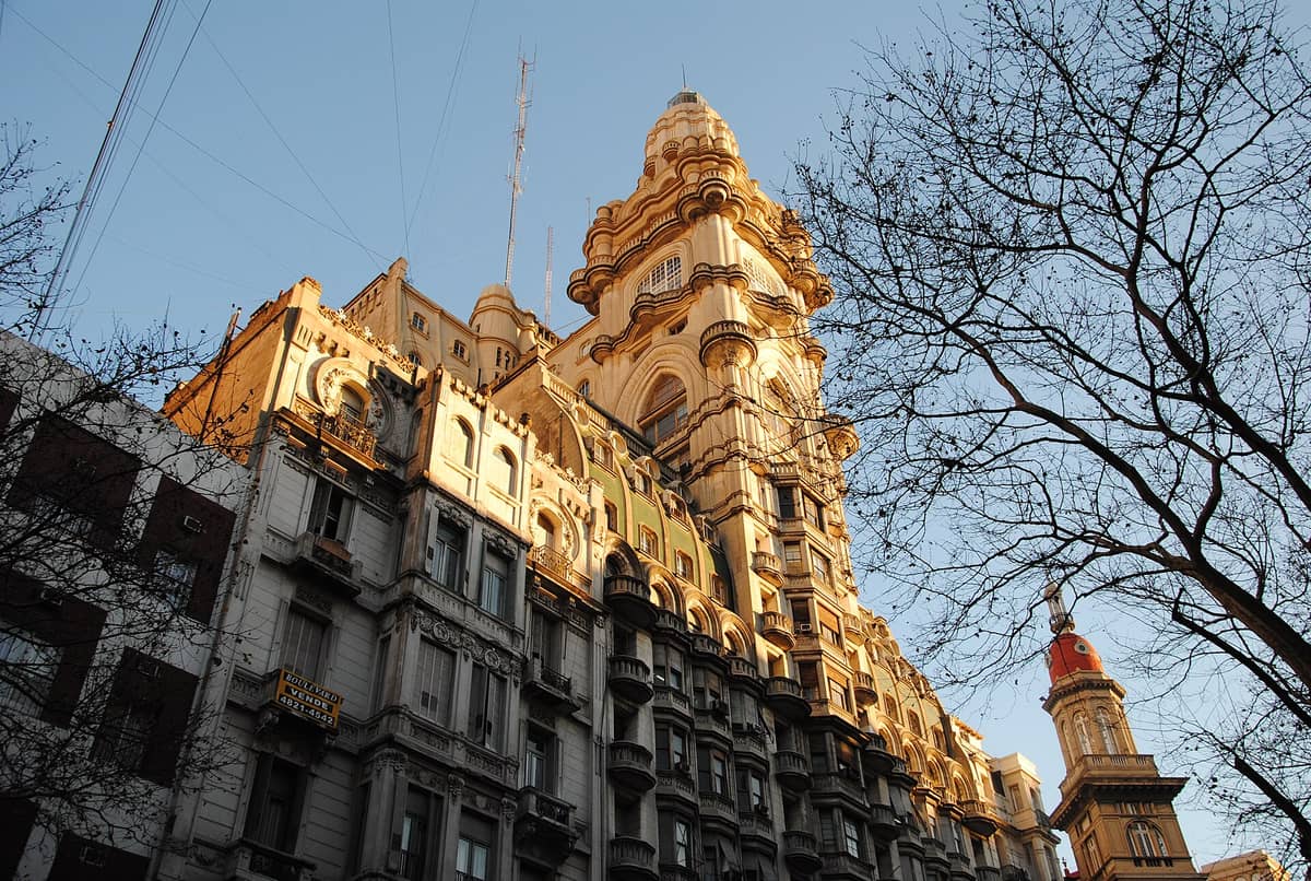 ภาพมุมกว้างของ Palacio Barolo โชว์ด้านหน้าตึกและหอคอยสุดแปลกตา แสงทองส่องโดน พร้อมกิ่งไม้ไร้ใบช่วยขับลายละเอียดทางสถาปัตยกรรม