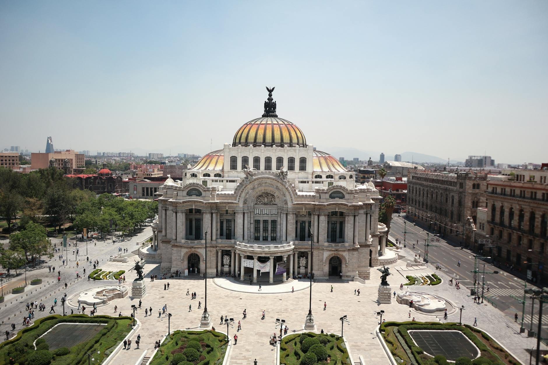 Vista ampia e soleggiata del Palacio de Bellas Artes a Città del Messico, con la sua iconica cupola, la facciata in marmo e i giardini circostanti animati dai visitatori.
