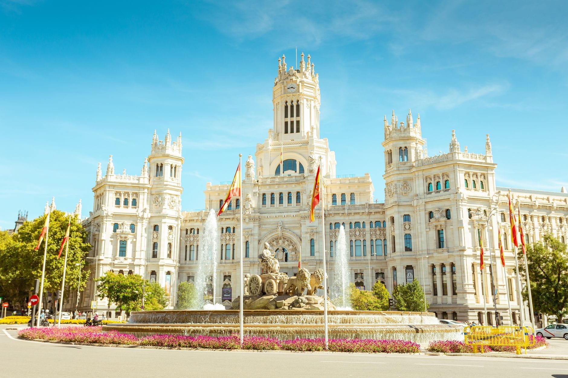 Il Palacio de Cibeles a Madrid in una giornata di sole, con un cielo azzurro intenso, bandiere spagnole e la celebre fontana di Cibeles in primo piano.
