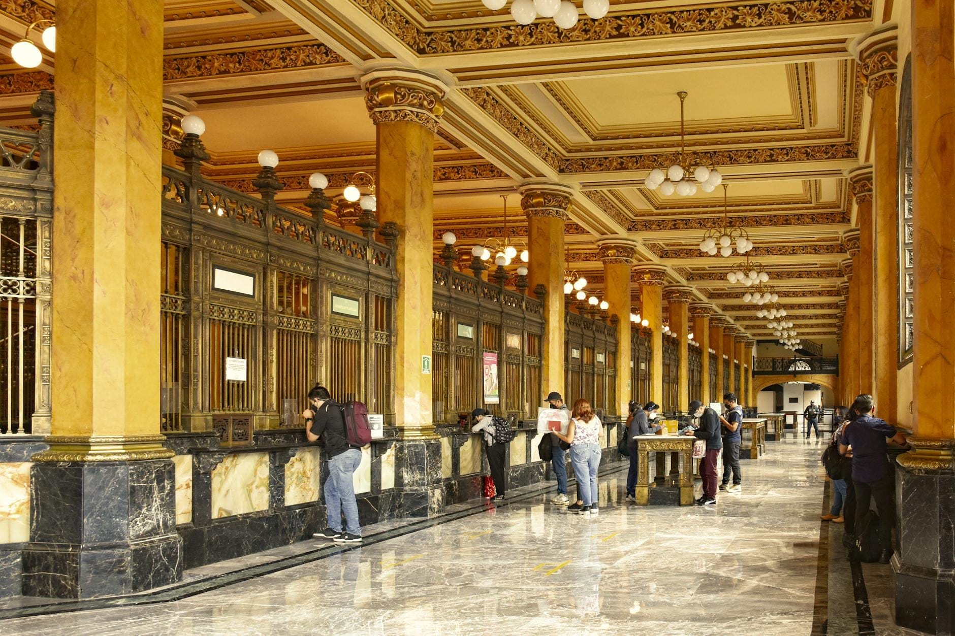 Visitatori agli eleganti banconi di marmo e oro all'interno del Palacio Postal di Città del Messico, con alte colonne dorate e soffitti riccamente decorati.