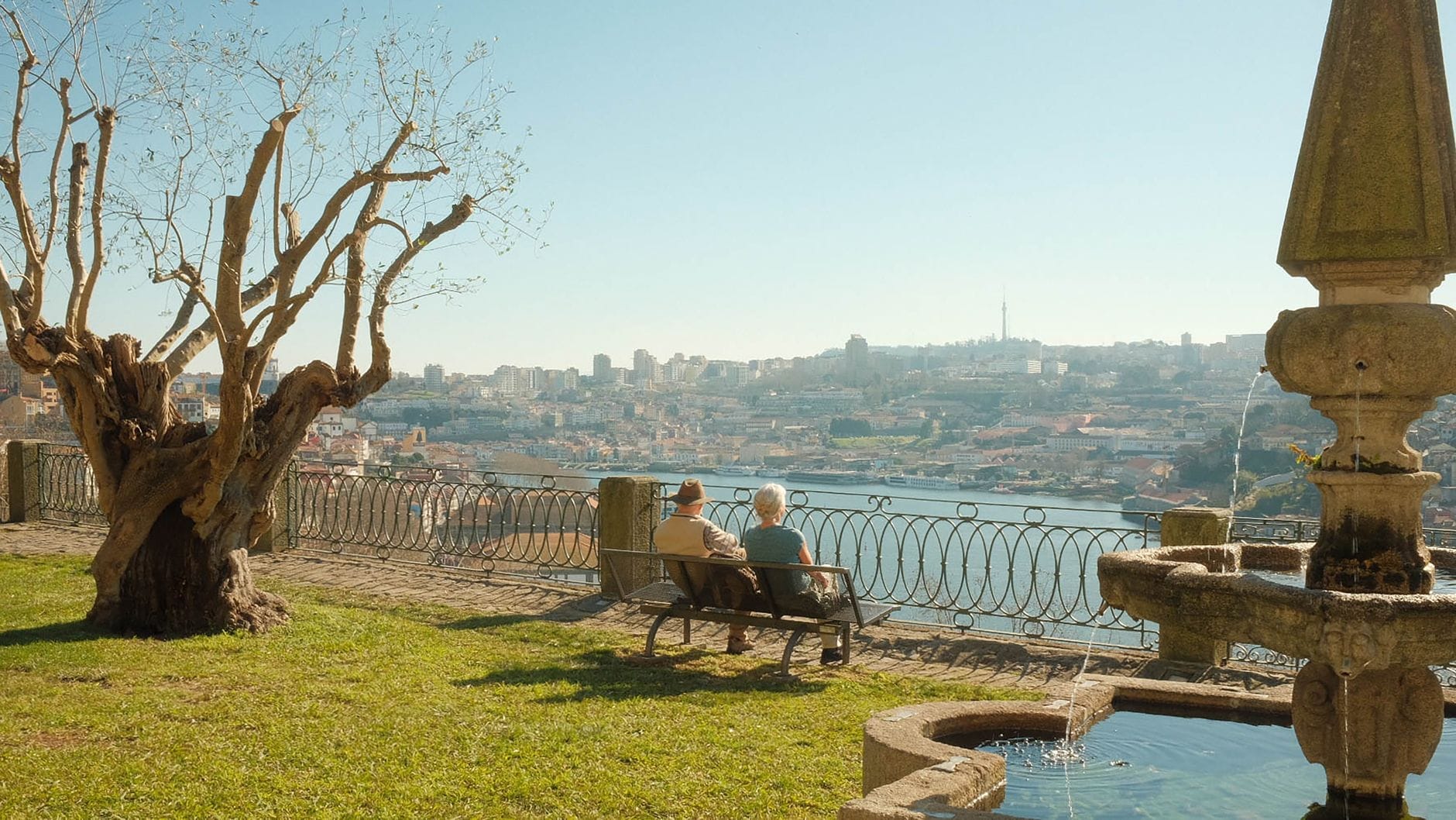 Due persone sedute su una panchina nei Jardins do Palácio de Cristal, con vista panoramica sul fiume Douro, Porto sullo sfondo e una classica fontana da giardino nelle vicinanze.