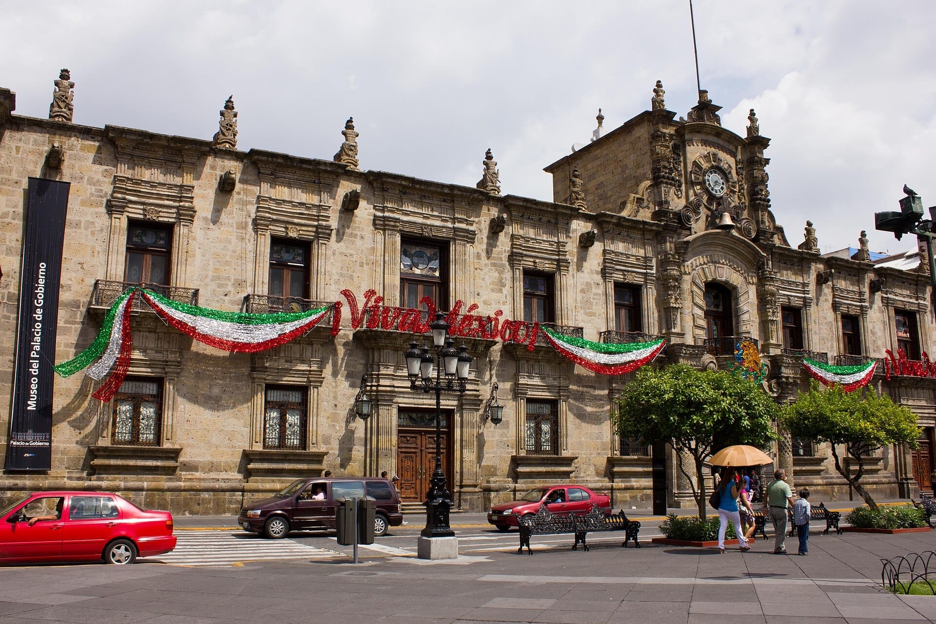 Esterno storico del Palacio de Gobierno de Jalisco adornato con festose bandiere messicane e l'insegna 'Viva México', con auto e pedoni davanti.