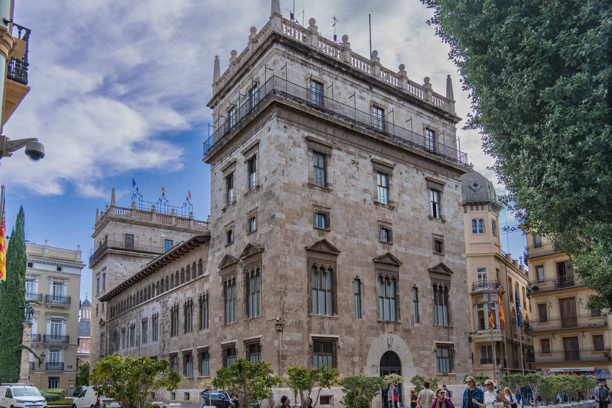 Vista ampia del Palacio de la Generalitat Valenciana, un grande edificio governativo in pietra tardo-gotica a Valencia, visto dalla strada con persone e bandiere visibili.