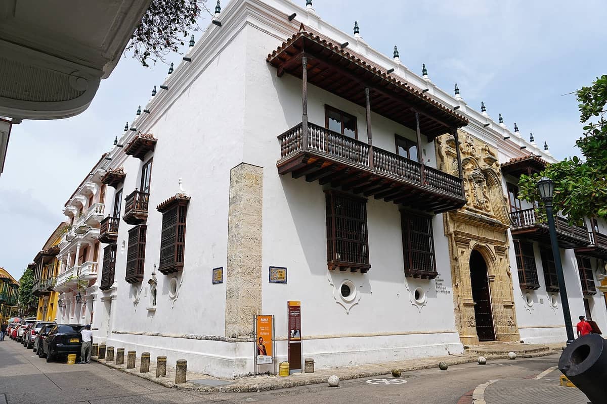 The Palacio de la Inquisición in Cartagena features a grand Baroque facade, whitewashed walls, wooden balconies, and ornate stone entrance under bright daylight.