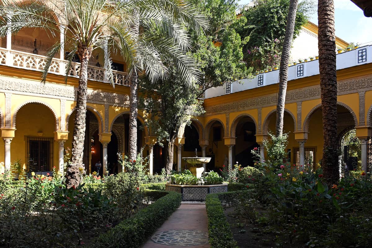 สวนใจกลางวังเขียวขจี มีต้นปาล์ม น้ำพุ และอาคารสองชั้นสีเหลืองอาร์เคด Palacio de las Dueñas เมืองเซบีญา