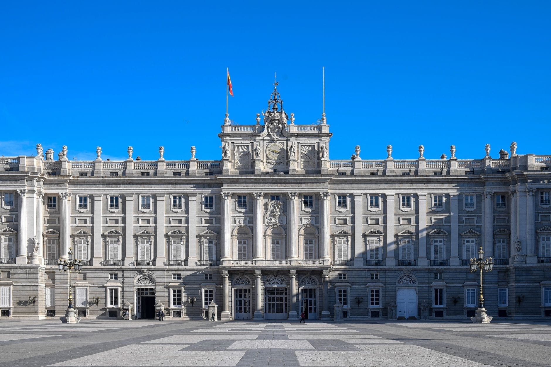 Facciata principale del Palazzo Reale di Madrid sotto un cielo azzurro limpido, con un'ampia piazza aperta e un'architettura simmetrica, perfetta come immagine di copertina per un'attrazione turistica.