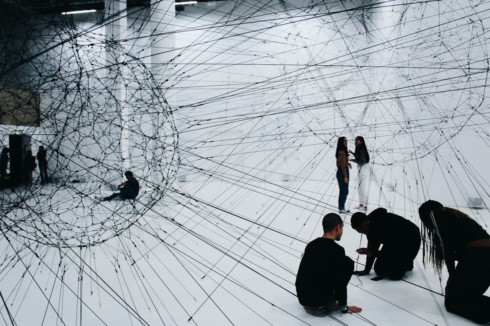 Visitatori che interagiscono con un'installazione d'arte contemporanea immersiva e di grande formato, realizzata con fili neri in uno spazio espositivo bianco.