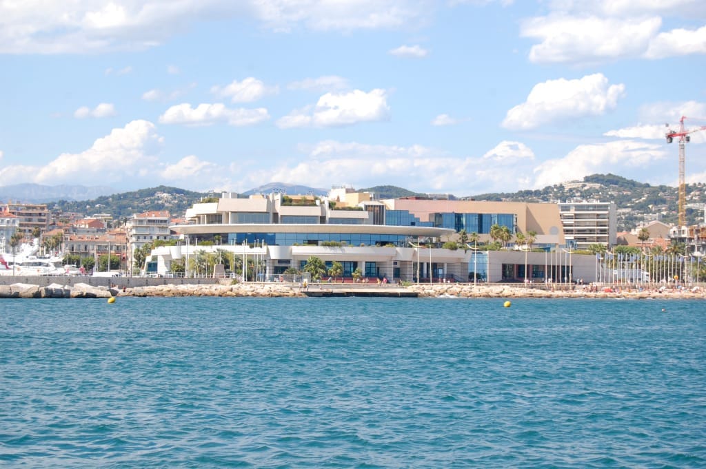 L'edificio del Palais des Festivals et des Congrès visto dall'acqua, con palme, cieli soleggiati e il panorama urbano di Cannes sullo sfondo.