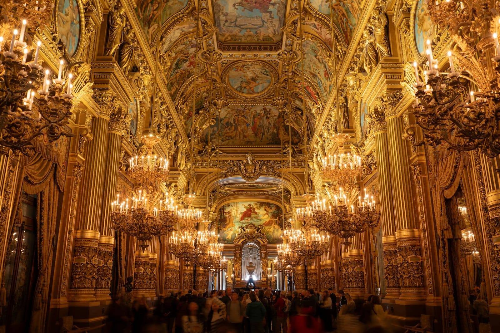 Il grande salone sontuosamente decorato del Palais Garnier, con ornamenti dorati, lampadari e un soffitto dipinto, animato dai visitatori che ammirano l'architettura.