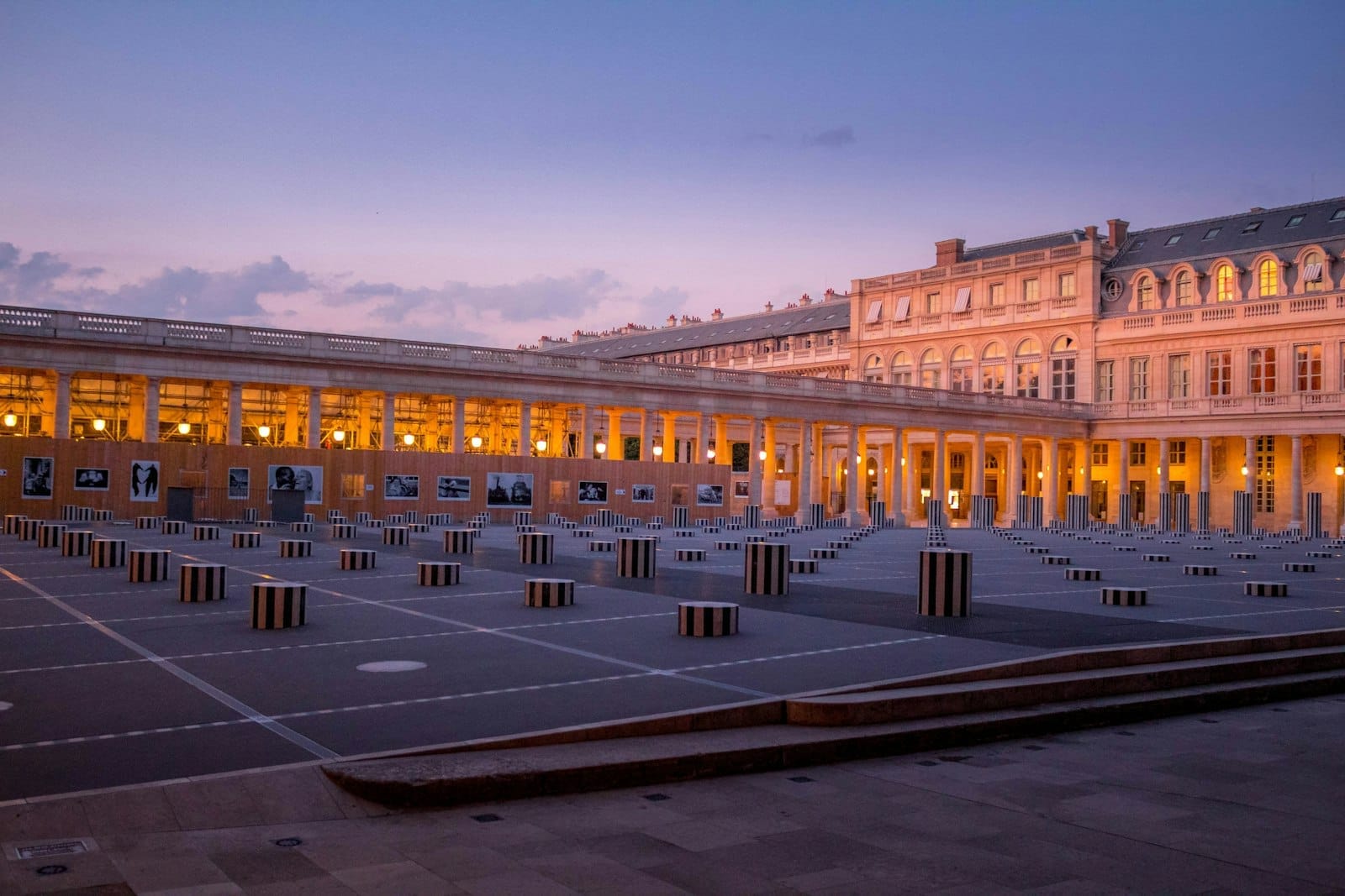 Il cortile del Palais Royal al tramonto con le iconiche colonne di Buren in bianco e nero, i portici classici e le luci calde dell'edificio sotto un cielo viola.