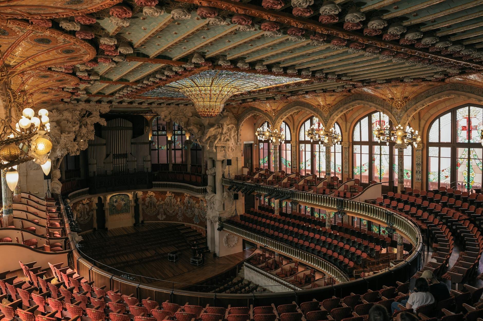 Wide-angle view of the Palau de la Música Catalana’s stunning main concert hall, featuring ornate stained glass ceiling, arched windows, beautiful balconies and rows of red seats.