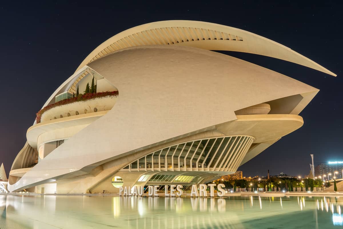Vista notturna del Palau de les Arts Reina Sofía a Valencia: l'iconica struttura bianca e curva si riflette nell'acqua accanto all'insegna illuminata.