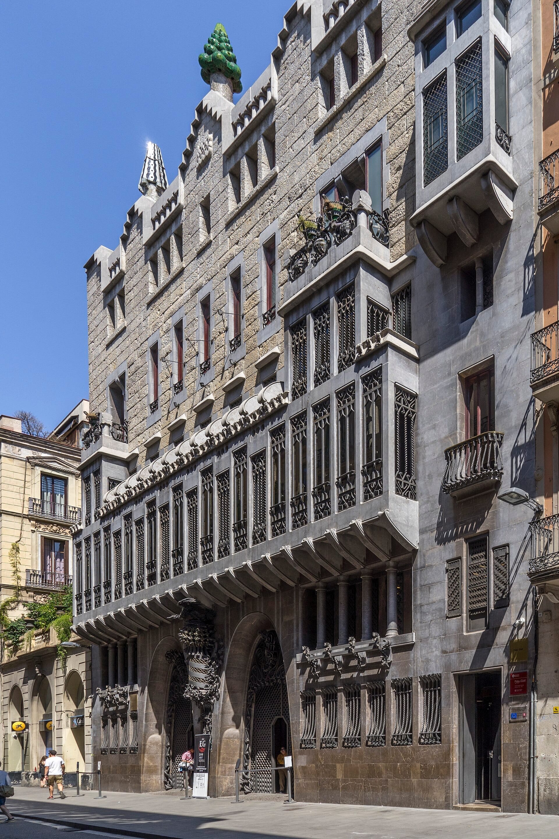 Facade of Palau Güell in Barcelona, featuring ornate wrought-iron details, unique window shapes, and colorful rooftop chimneys under a clear blue sky.