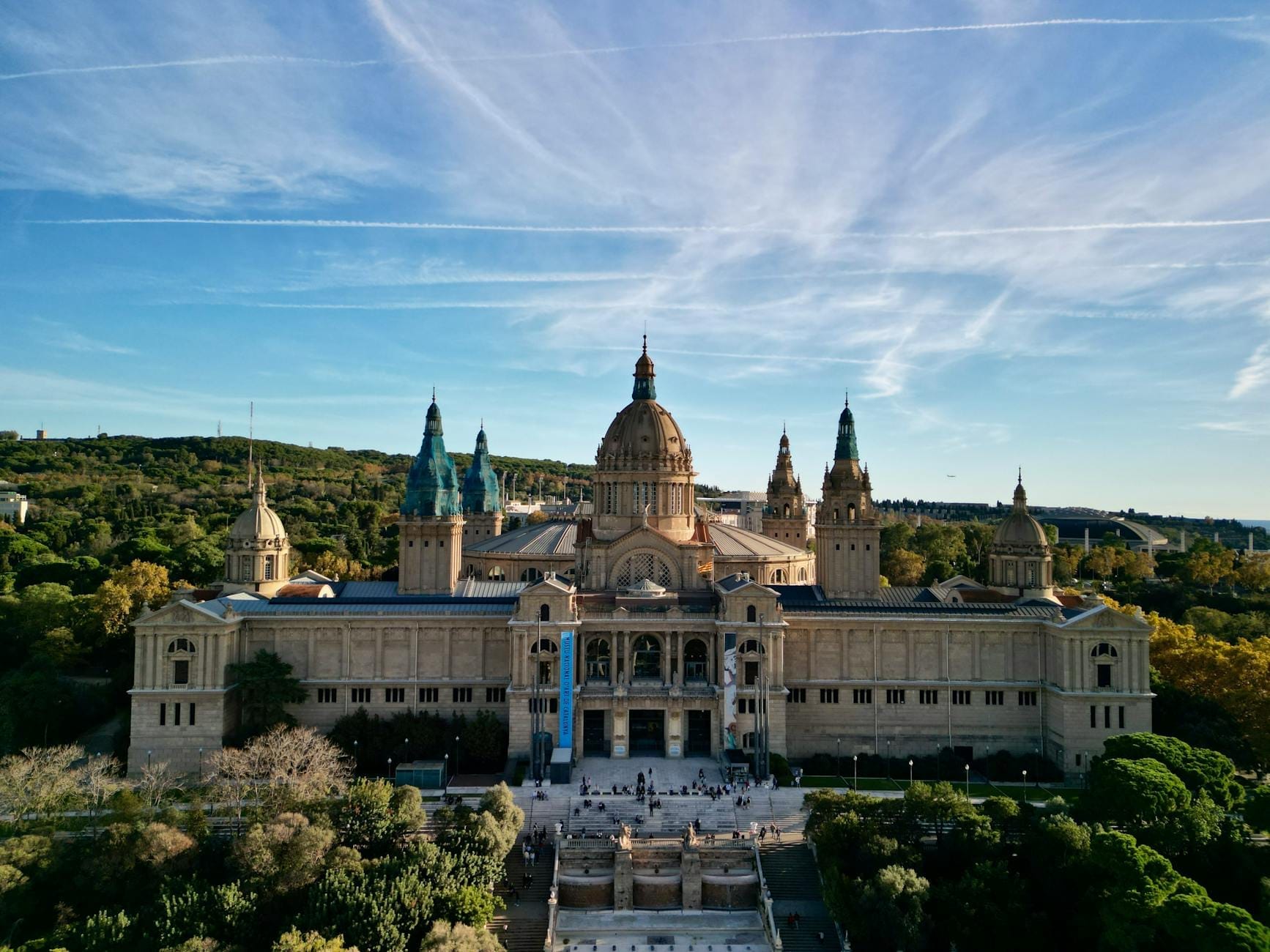 A stunning wide-angle aerial view of the Palau Nacional atop Montjuïc, surrounded by green trees with blue sky and wispy clouds above, in Barcelona.