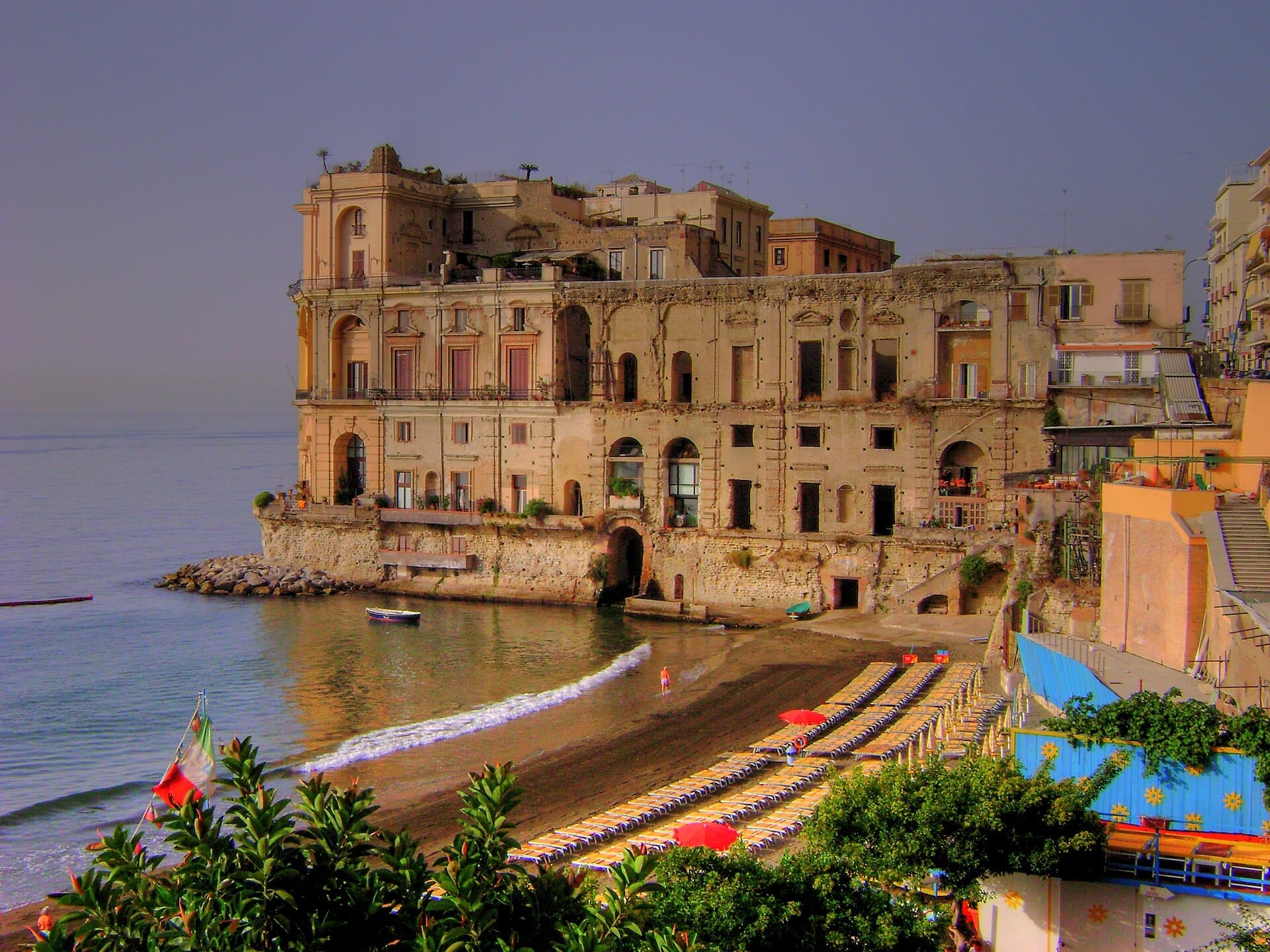 La facciata incompiuta del Palazzo Donn'Anna si erge sul mare, affacciata su una spiaggia di sabbia fiancheggiata da lettini vuoti e ombrelloni colorati.