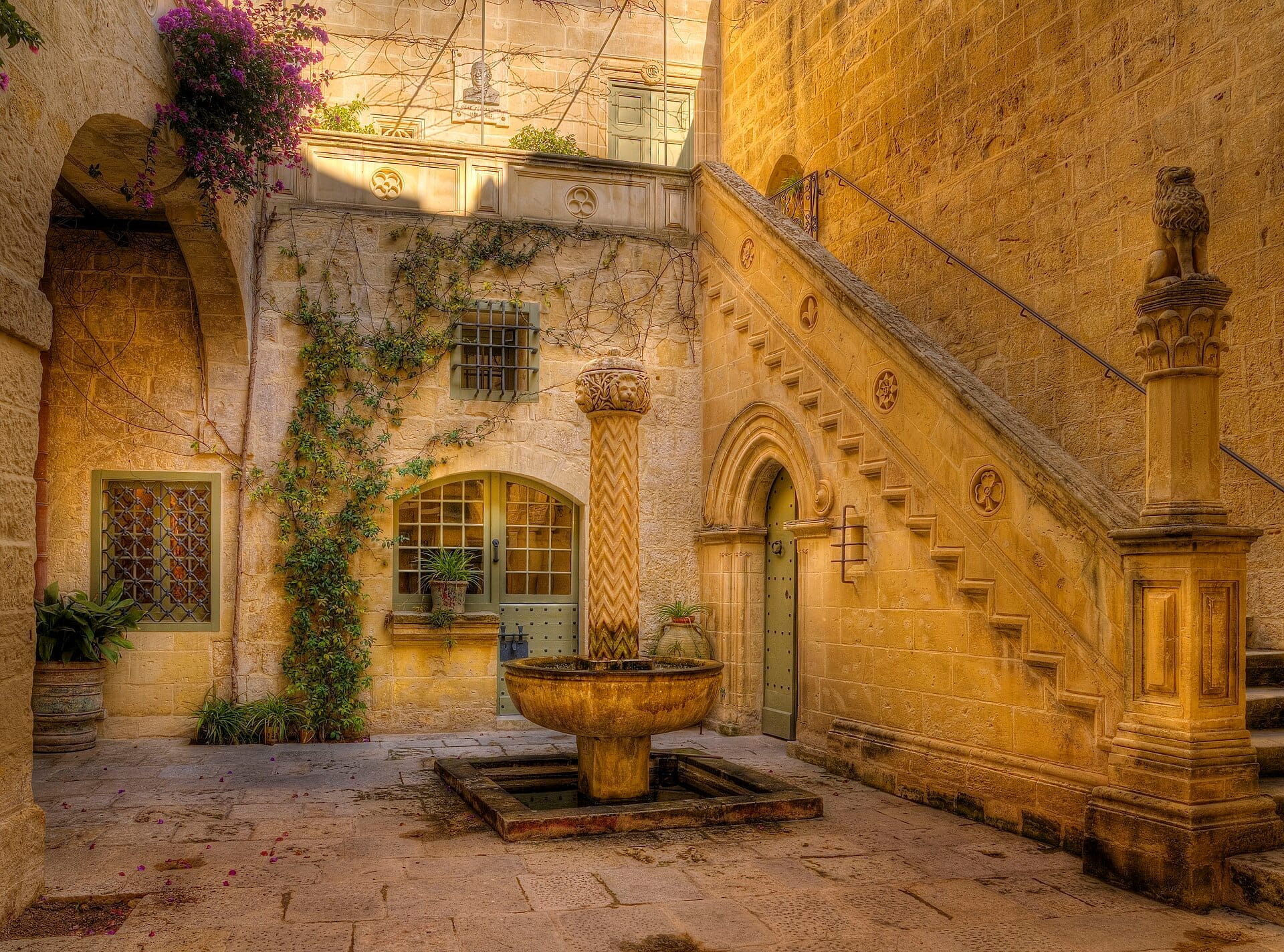 Cortile in pietra al Palazzo Falson Historic House Museum con una fontana centrale, una scalinata ad arco, piante fiorite e una calda luce dorata del sole.