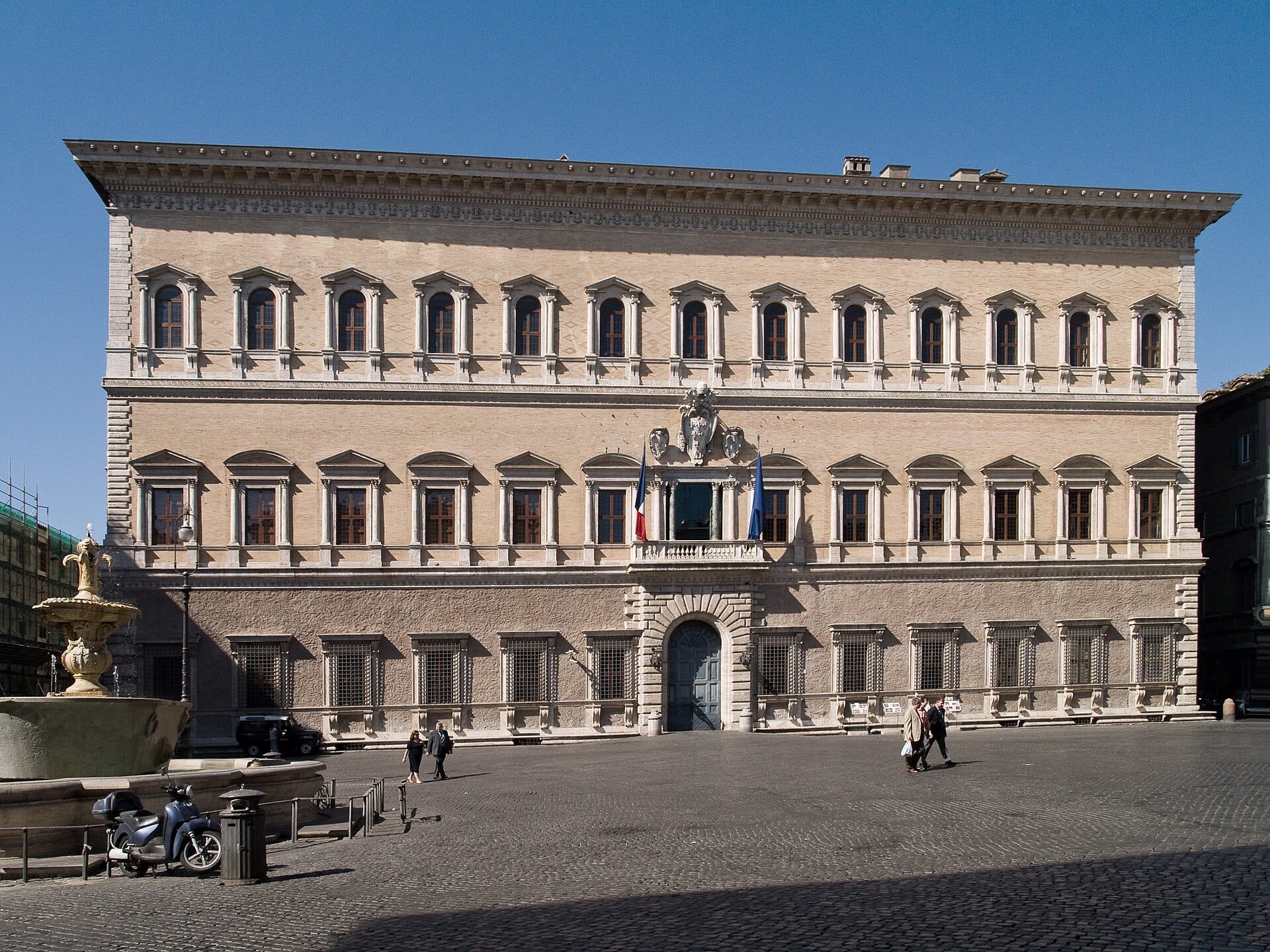 Front view of Palazzo Farnese in Rome, showing Renaissance architecture, symmetrical windows, French flags, and people walking in the sunny piazza.