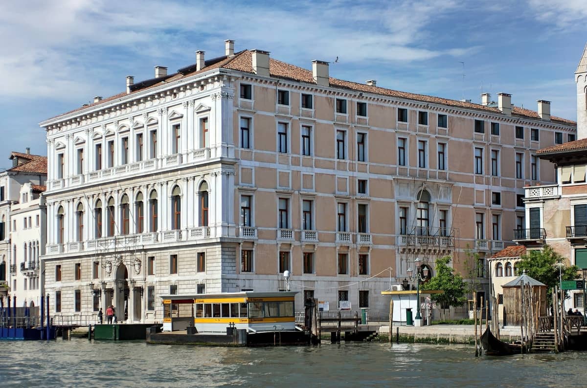 Die neoklassische Fassade des Palazzo Grassi, vom gegenüberliegenden Ufer des Canal Grande aus gesehen, mit eleganten Fenstern und einer Vaporetto-Haltestelle davor.