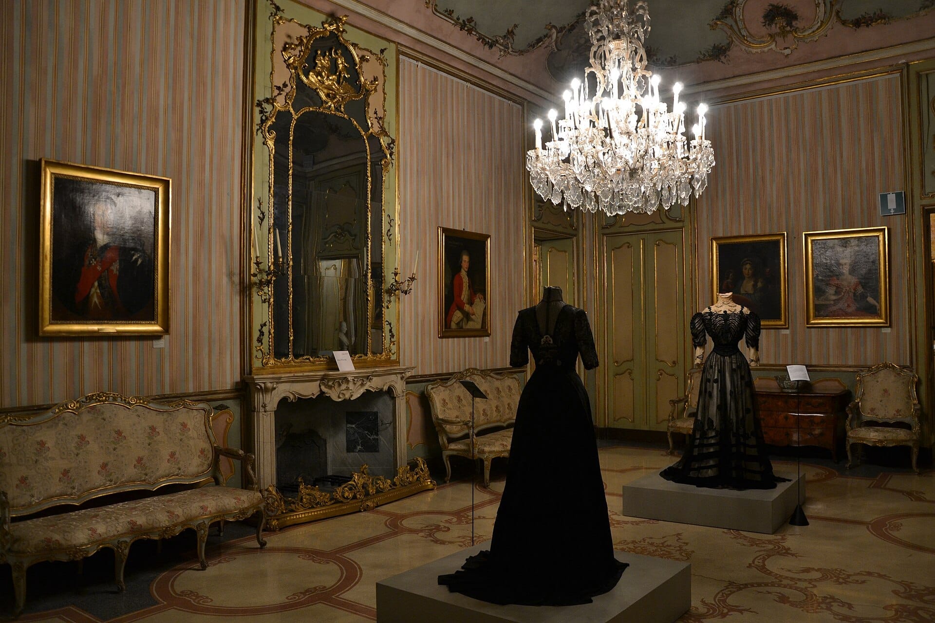 Due elaborati abiti neri in esposizione in un sontuoso salone di Palazzo Morando, con divani antichi, specchio dorato, dipinti e un lucente lampadario.