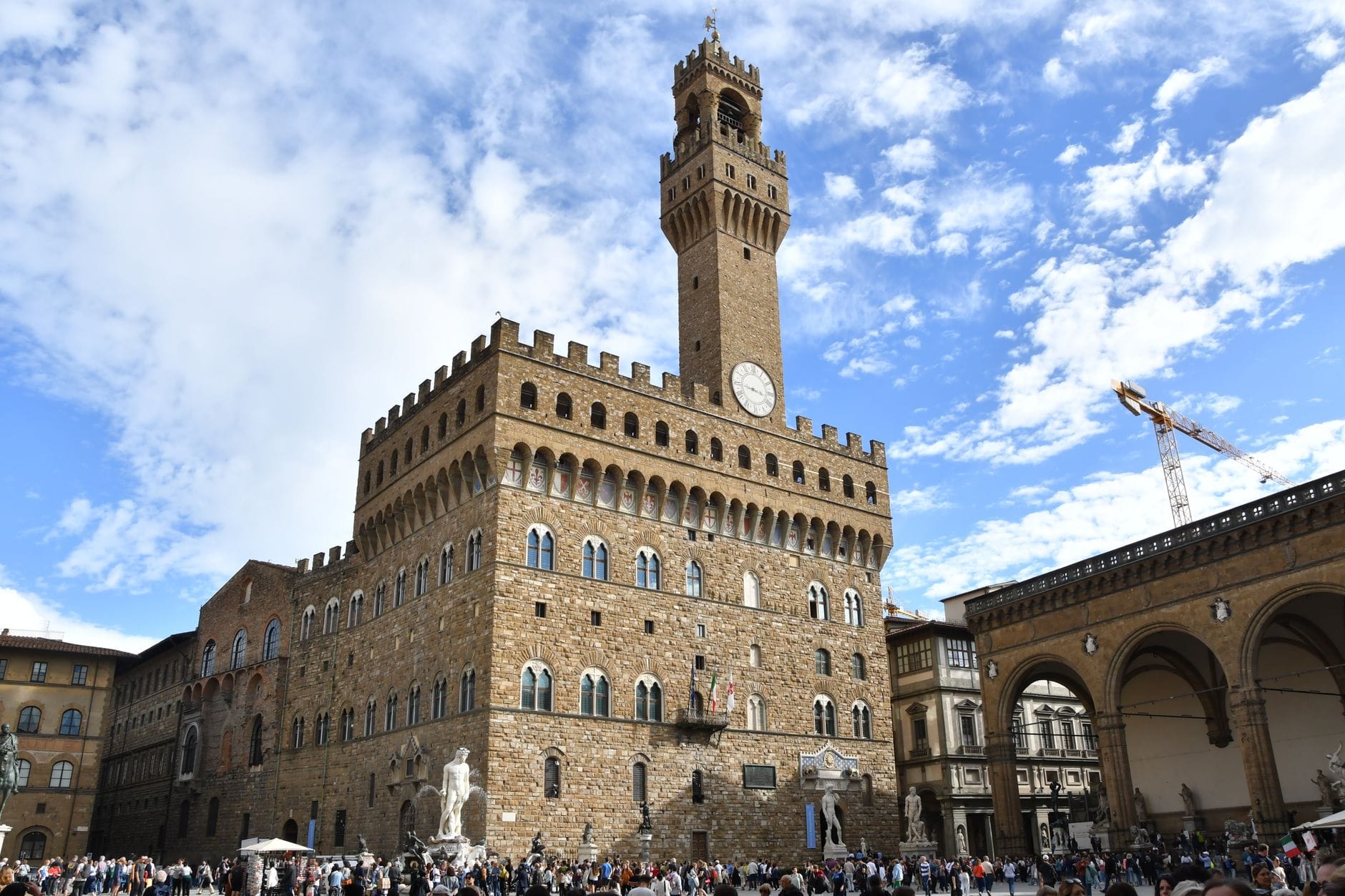 วิวกว้างด้านหน้าของ Palazzo Vecchio กับหอคอย Arnolfo ที่เด่นตระหง่านเหนือ Piazza della Signoria อันวุ่นวายในฟลอเรนซ์ ท่ามกลางท้องฟ้าสีฟ้าปนเมฆขาว