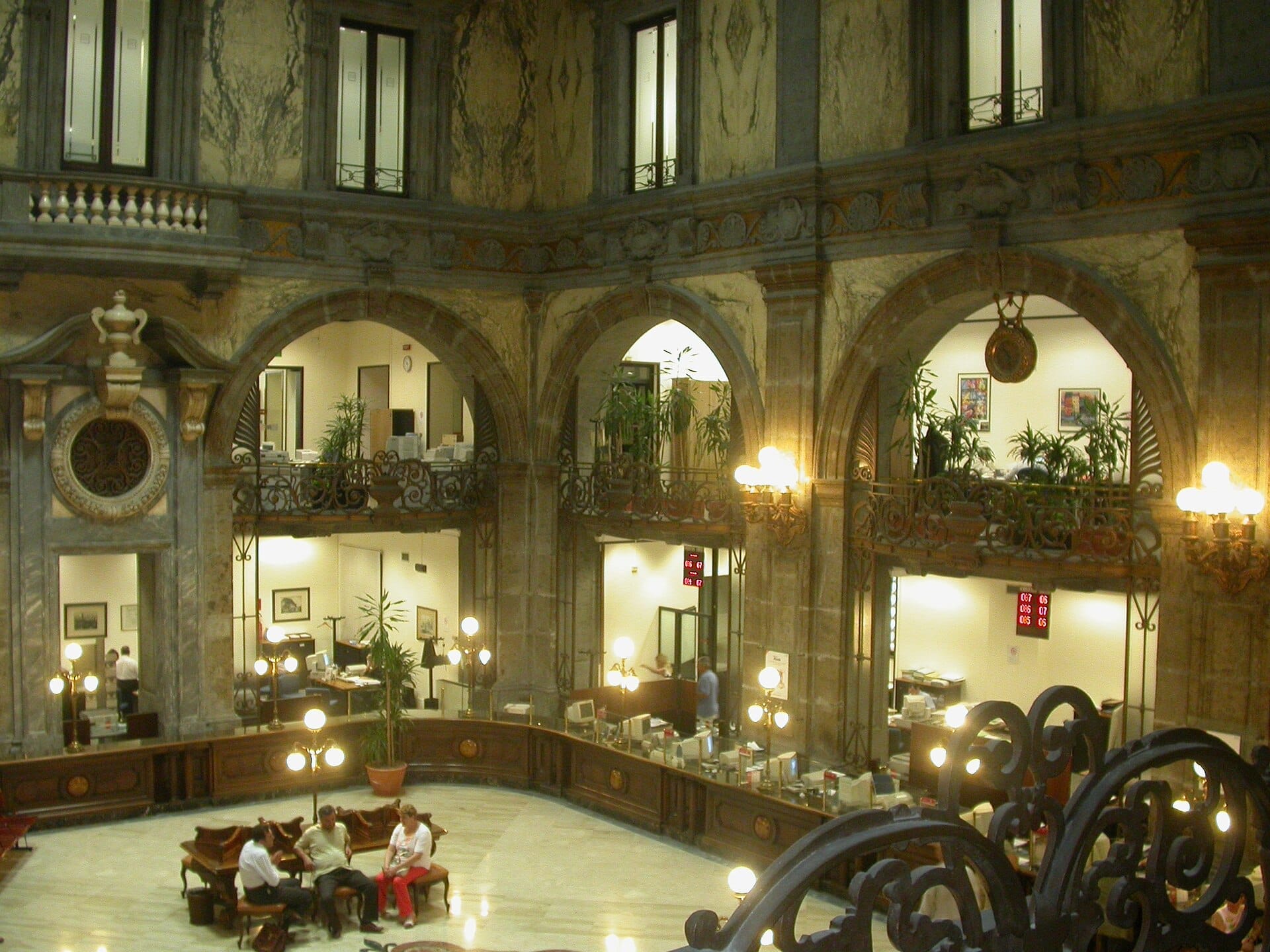 Ampia sala barocca all'interno del Palazzo Zevallos Stigliano, con archi in marmo, ringhiere ornate e persone sedute sotto lampadari luminosi.