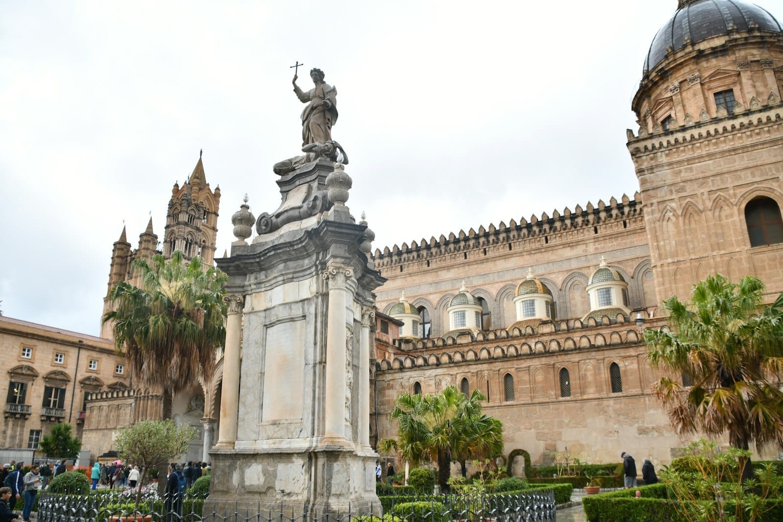 Statue und Palmen vor der prachtvollen Kathedrale von Palermo; Menschen schlendern bei bewölktem Himmel über den Platz.