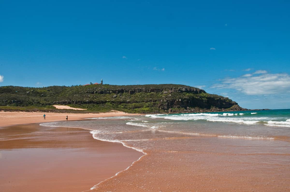 Palm Beach, Sídney, con el icónico faro de 1881 en lo alto del promontorio, arena dorada, olas suaves y cielo azul despejado en un día soleado.