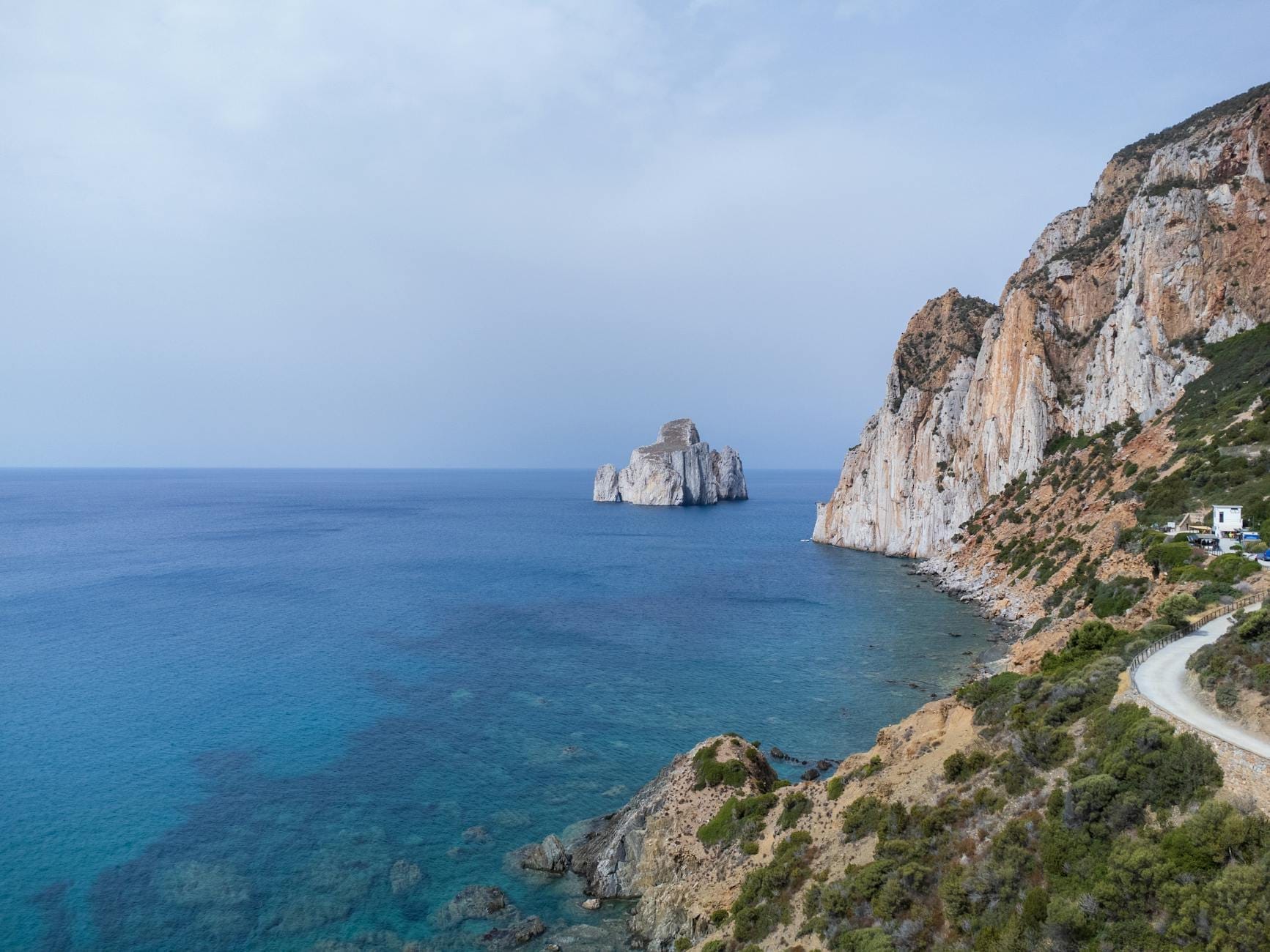 Weitwinkelperspektive auf den Meeresturm Pan di Zucchero, der sich aus dem türkisfarbenen Wasser neben dramatischen Felsküsten an Sardiniens Südwestküste erhebt – aufgenommen unter einem strahlend klaren Himmel.