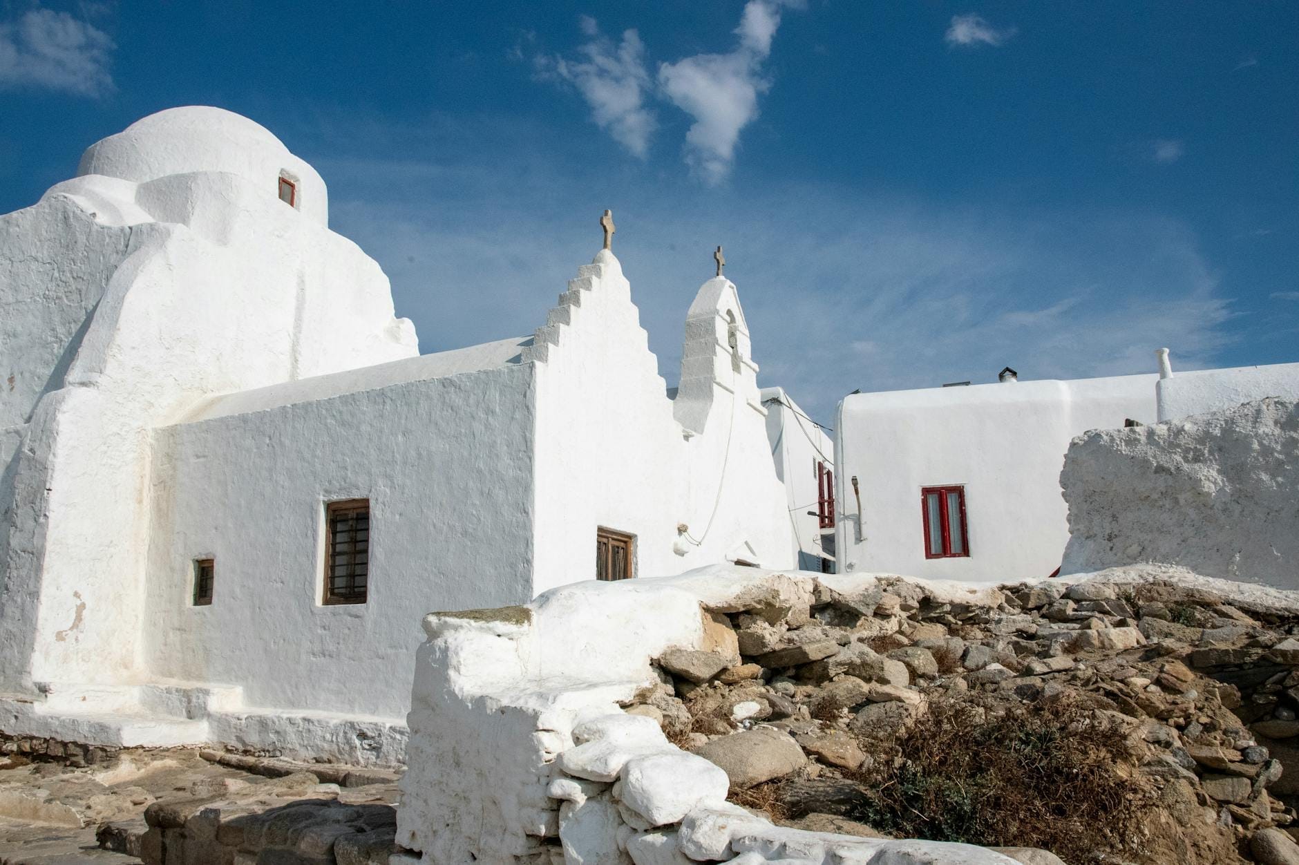 Vista amplia del exterior encalado y asimétrico de la Panagia Paraportiani con sus formas arquitectónicas únicas bajo un cielo azul intenso en Mykonos, Grecia.