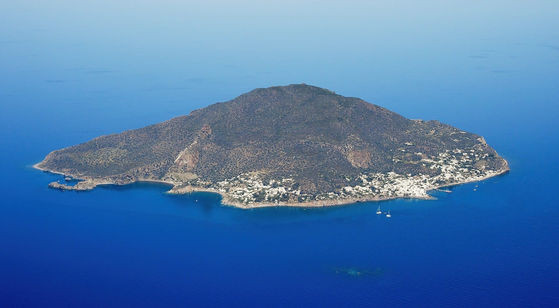 Luftaufnahme der Insel Panarea mit zerklüftetem Vulkangelände, weiß getünchten Gebäuden und dem tiefen blauen Tyrrhenischen Meer rund um die Küste.