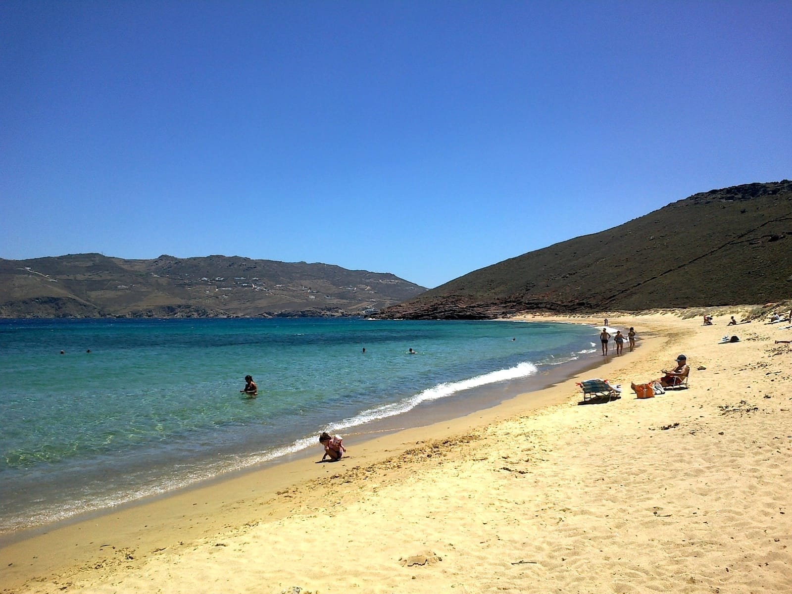 Arena dorada y aguas cristalinas de color azul se curvan a lo largo de Panormos Beach, con algunos bañistas y nadadores frente a un fondo de colinas rocosas.
