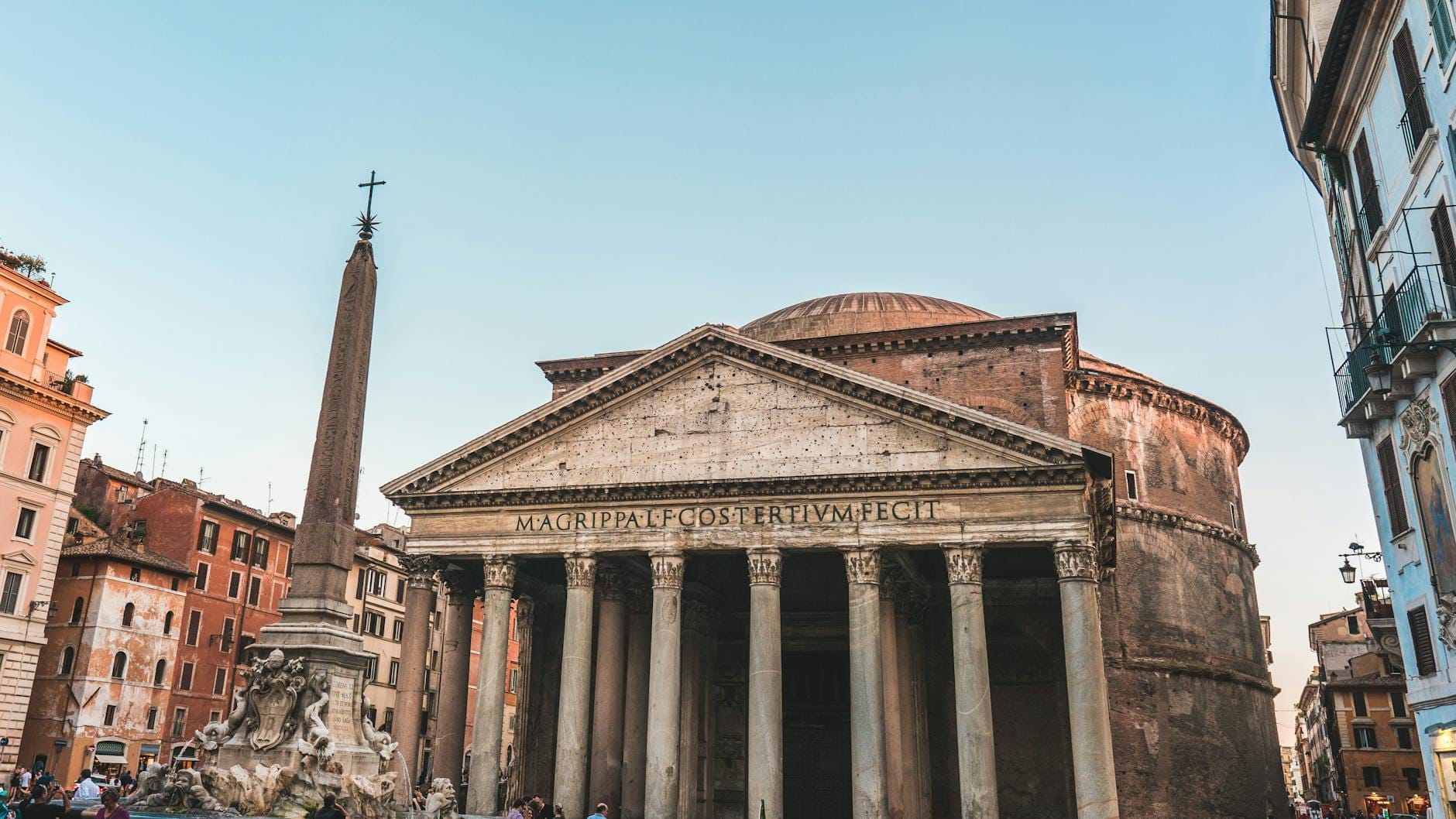 Wide view of the Pantheon's grand facade and dome with its iconic columns and nearby fountain, surrounded by lively Rome city buildings under a clear sky.